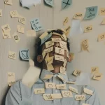 Male college student standing in a corner with blue and yellow sticky notes all over the wall and his face and body. He is trying to manage stress while in college.