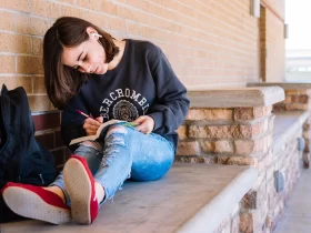 female community college student sitting outside with her backpack beside her writing in a notebook