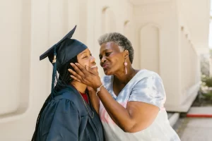 Female wearing a black graduation gown being kissed on the cheek by a family member. This shows family support.