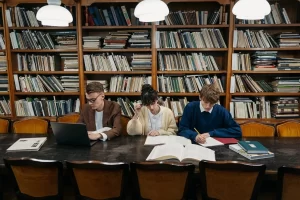 Three students sitting at a big black desk in a library studying to get good grades