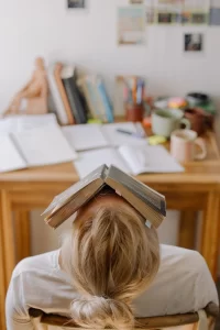 female student sitting in front of a desk. There is a book one her face. She is exhausted as she studies to get good grades