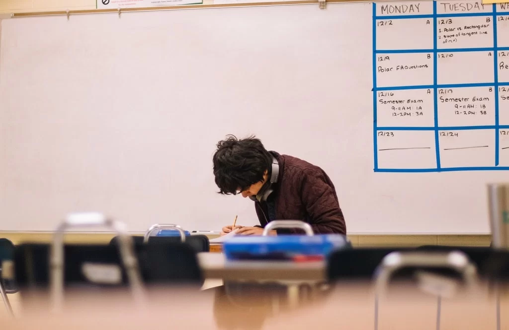 Dual enrollment high school student sitting in class with headphones around his neck writing at a desk.