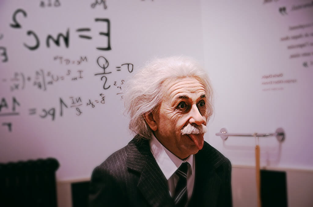 Albert Einstein in a black suit sticking his tongue out. A whiteboard of equations is blurred in the background