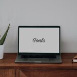 Laptop with the word goals in the screen is sitting on a brown desk with a plant and notebook beside it