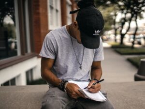 male college student sitting outside in a black ballcap writing in a notebook
