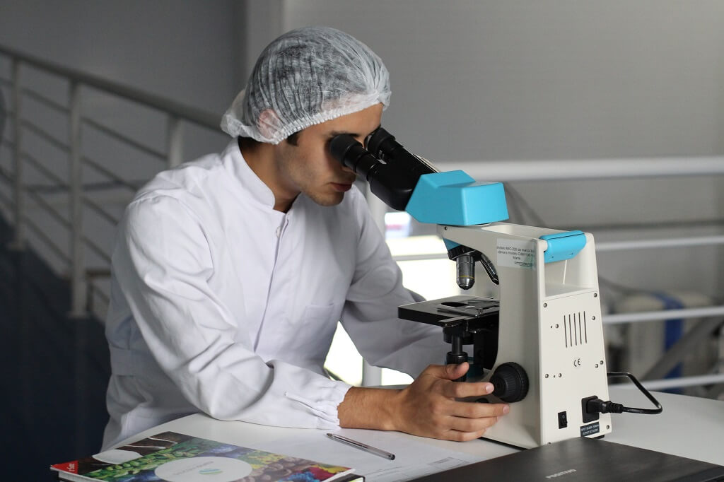 a man in a white lab coat is sitting at a table looking through a microscope. He has one of the highest-paying jobs in the medical field.